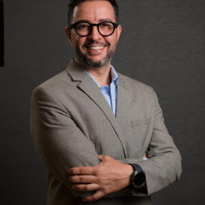 Adriano é um homem branco, de cabelos castanhos escuros e curtos, usa barba e óculos. Veste blazer cinza sobre camisa social azul claro