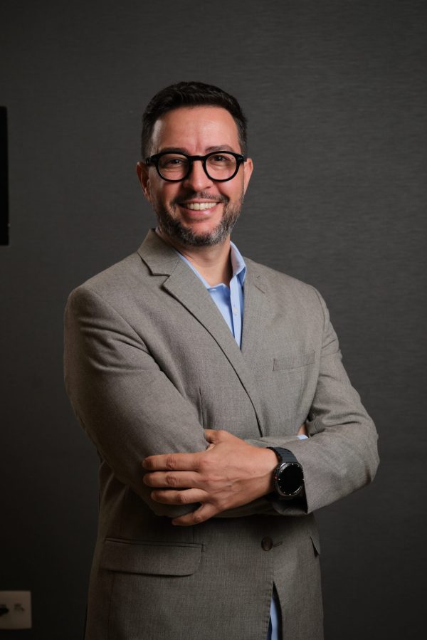 Adriano é um homem branco, de cabelos castanhos escuros e curtos, usa barba e óculos. Veste blazer cinza sobre camisa social azul claro