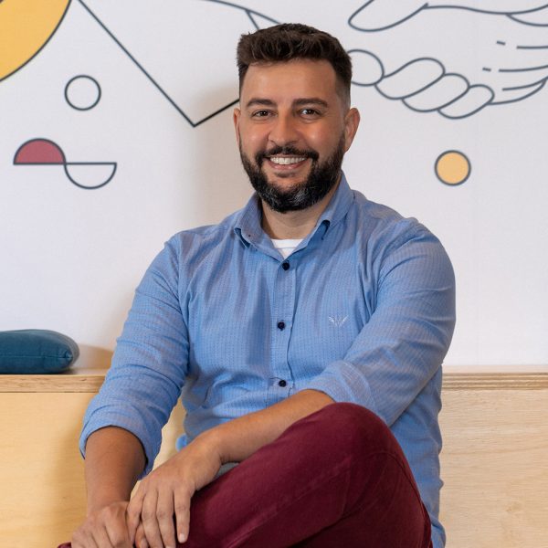 Adriano é um homem branco, de cabelos castanhos escuros e curtos, usa barba. Veste camisa social azul claro sobre camiseta branca com calças em tom vermelho